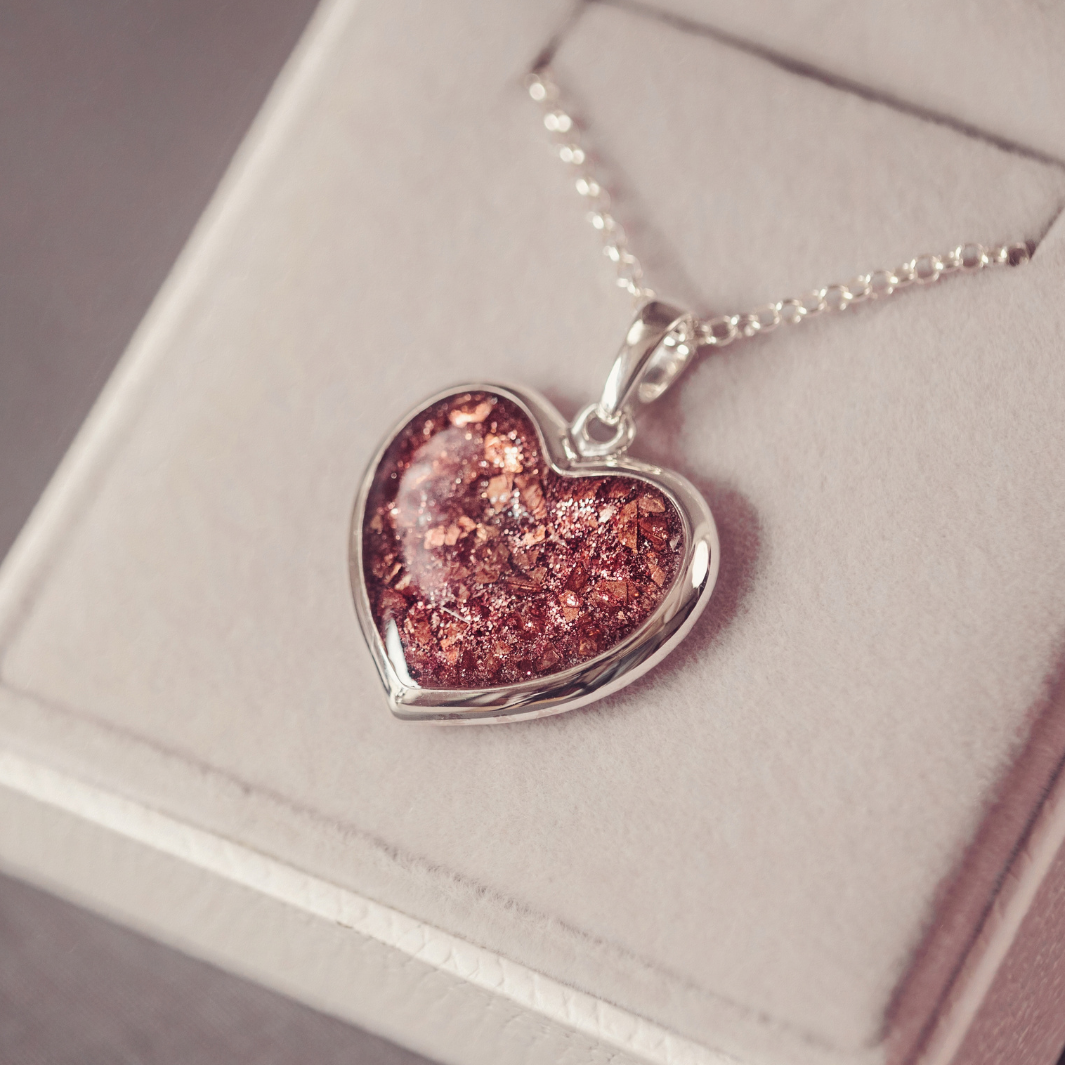 A heart-shaped pendant necklace with a sparkling coppery brown glitter center and a silver-toned bezel, resting on a light beige jewelry box.