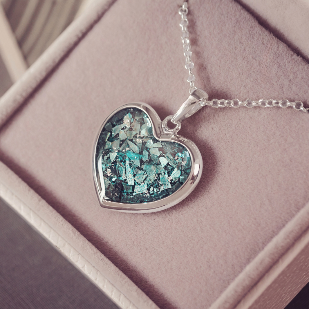 A silver heart pendant necklace with a textured, iridescent blue and green glitter-filled center, resting on a dusty pink jewelry box.
