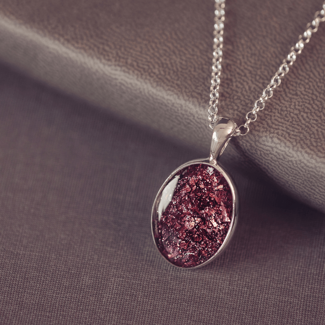 Sterling silver oval necklace with a central section filled with memorial ashes and brown crushed crystals, displayed on a grey jewellery box