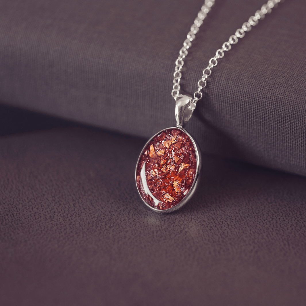 Sterling silver oval necklace with a central section filled with memorial ashes and orange crushed crystals, displayed on a dark background