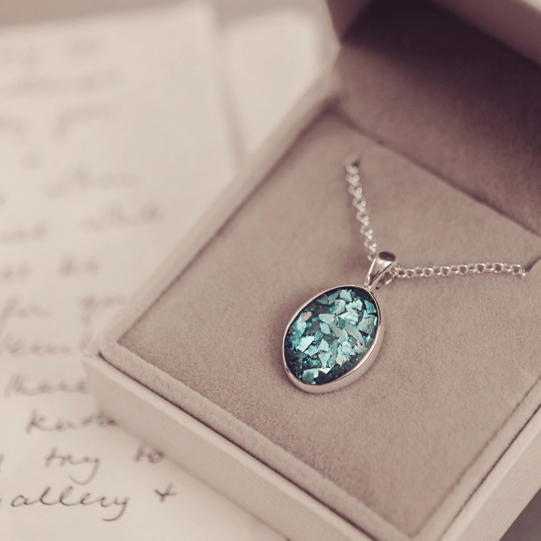 Sterling silver oval necklace with a central section filled with memorial ashes and turquoise crushed crystals, displayed on a grey fabric background