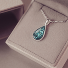A silver teardrop pendant necklace with a textured, glittery teal-blue center, resting on a beige jewelry box.