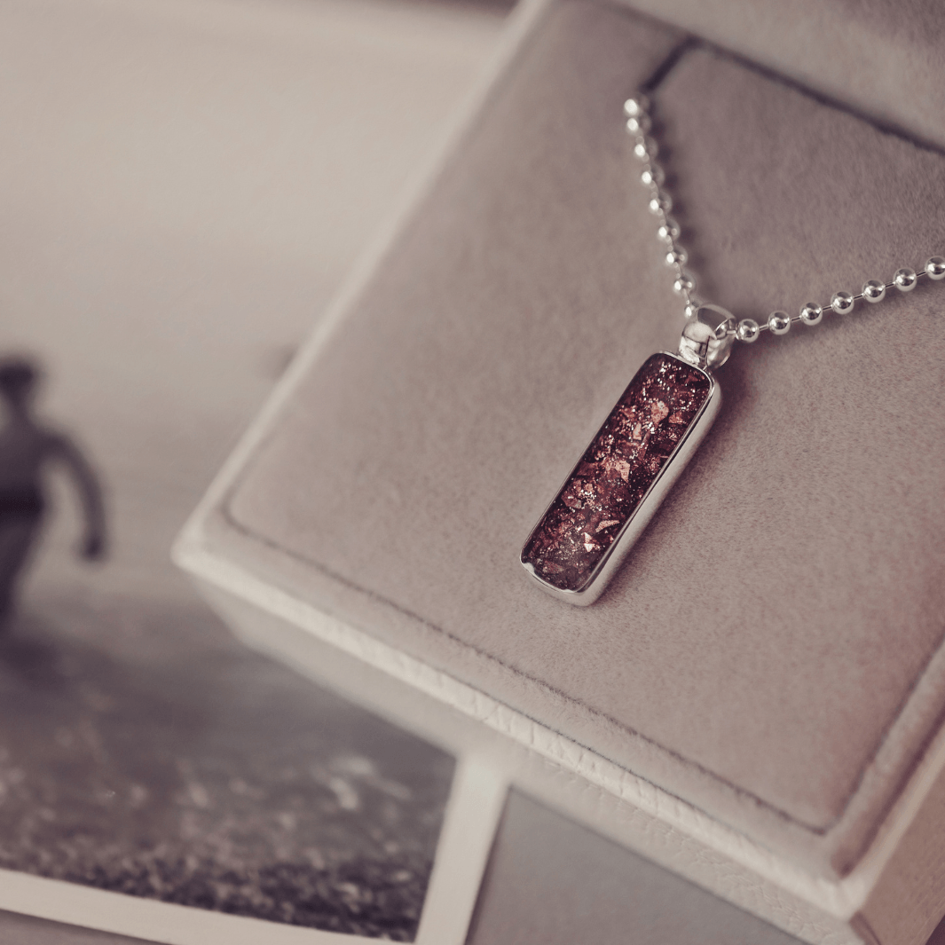 A sophisticated Mens silver necklace featuring a rectangular pendant filled with shimmering brown crystal and resin, delicately layered with cremation ashes, showcased against a dark background on a silver ball chain