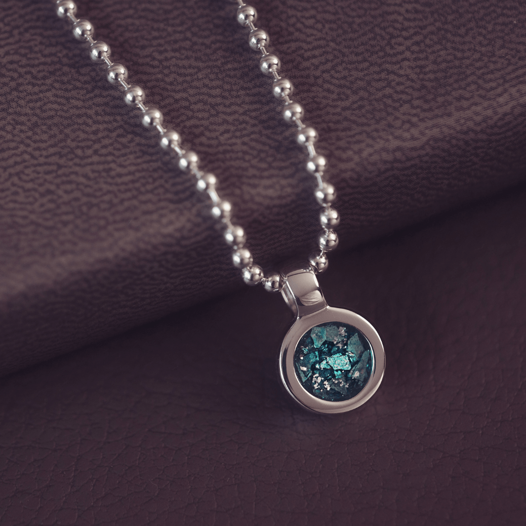 A sophisticated Mens silver necklace featuring a small round pendant filled with shimmering turquoise crystal and resin, delicately layered with cremation ashes, showcased against a dark background on a silver ball chain