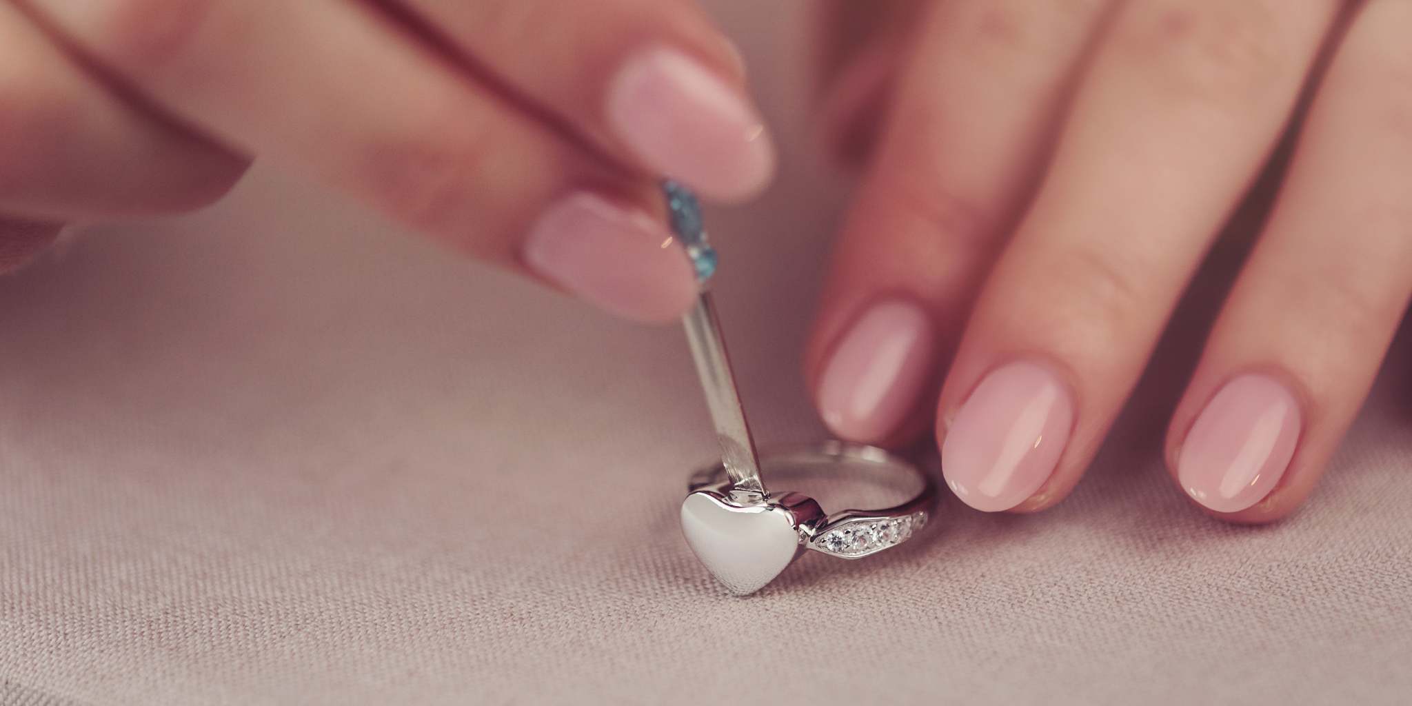 Close-up of hands with pink nail polish holding a silver ring with a heart design on a light background.