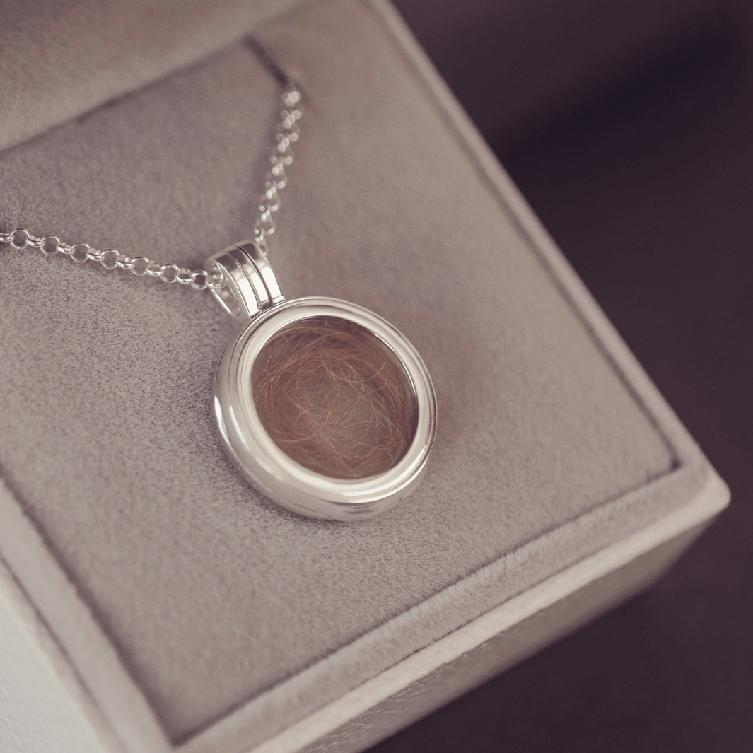 Round silver locket necklace with acrylic showing a locket of hair inside on a textured surface