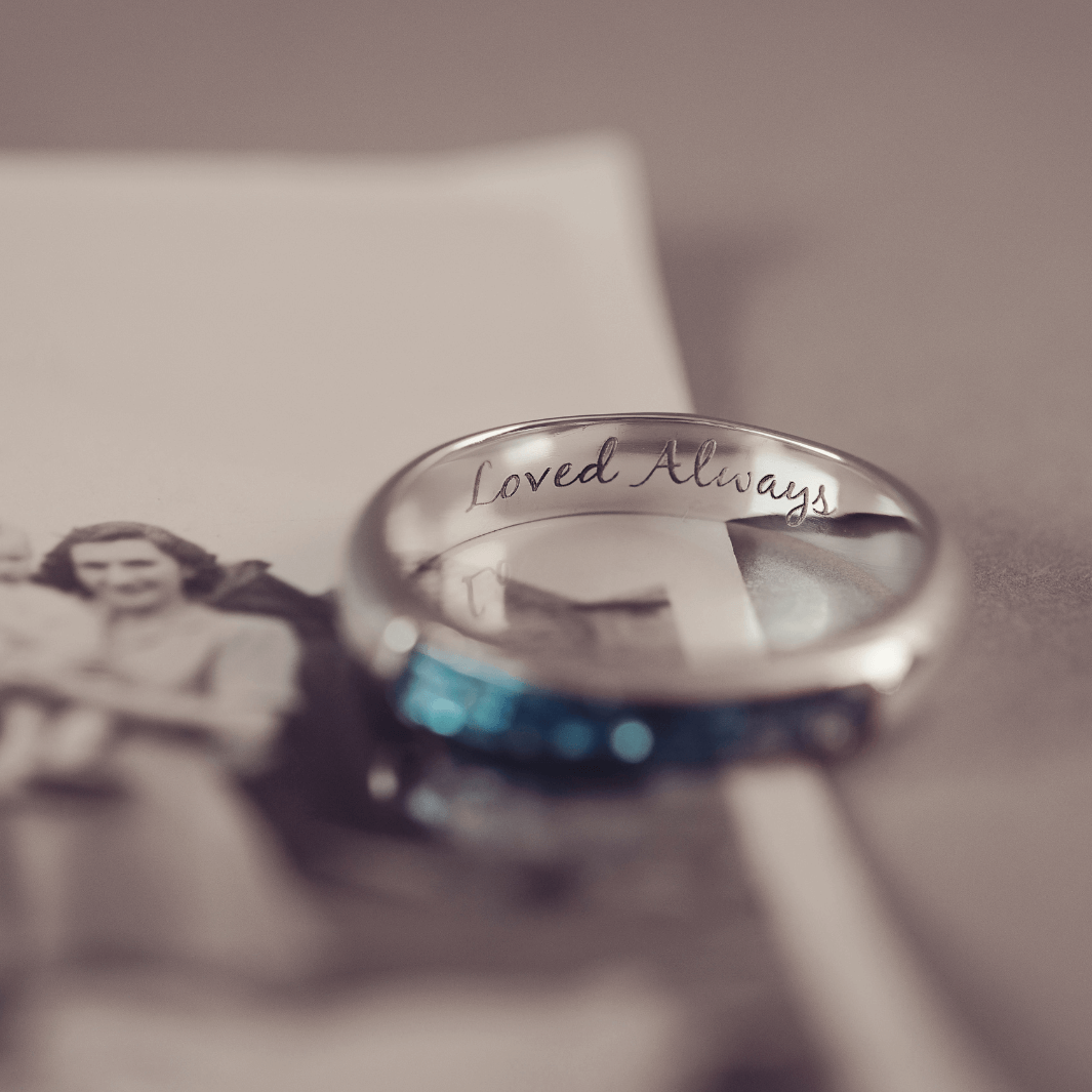 Sterling silver womens ring band with a central section filled with  memorial ashes and blue crushed crystals, on a blurred pale background. The ring has the message 'Loved Always' engraved into the inside of the ring band in black