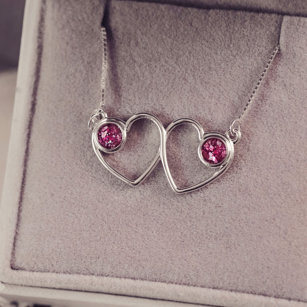 A delicate silver necklace featuring two intertwined heart-shaped pendants, each with a pink resin stone which holds crystal and cremation ashes. Displayed against a neutral background.