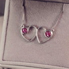A delicate silver necklace featuring two intertwined heart-shaped pendants, each with a pink resin stone which holds crystal and cremation ashes. Displayed against a neutral background.