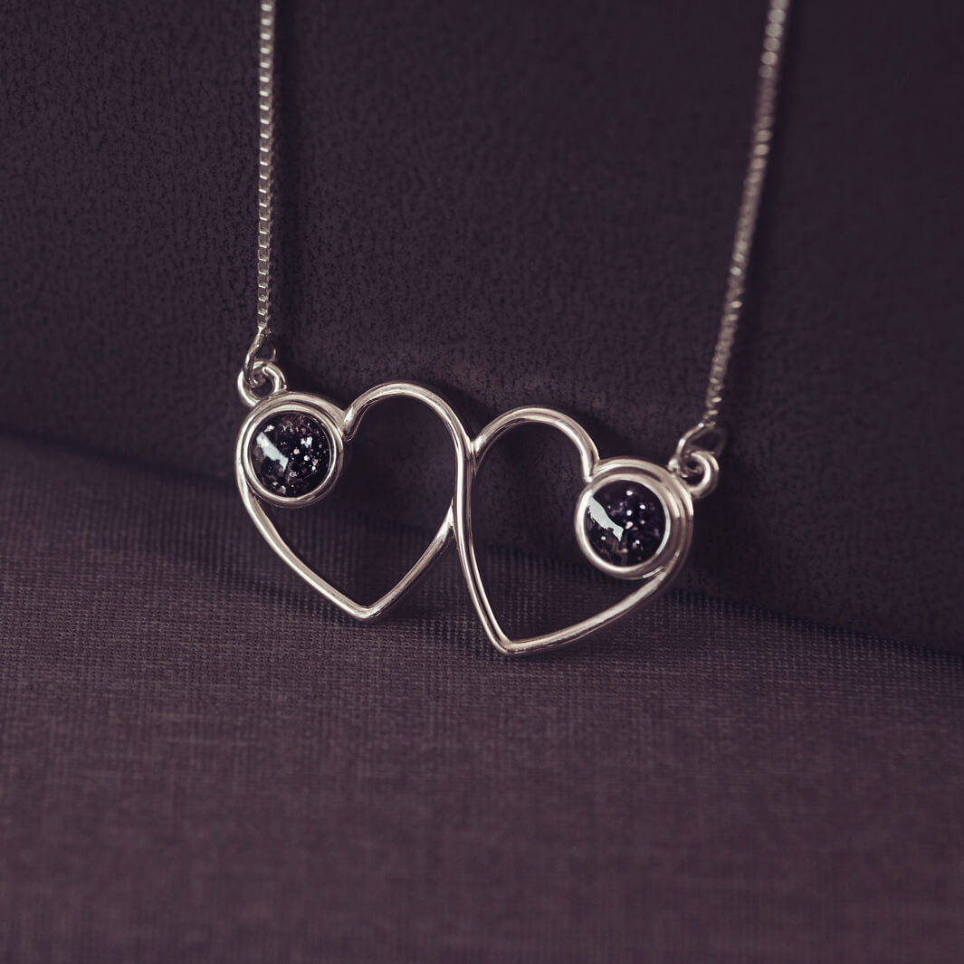 A delicate silver necklace featuring two intertwined heart-shaped pendants, each with a black resin stone which holds crystal and cremation ashes. Displayed against a neutral background.