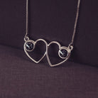 A delicate silver necklace featuring two intertwined heart-shaped pendants, each with a black resin stone which holds crystal and cremation ashes. Displayed against a neutral background.