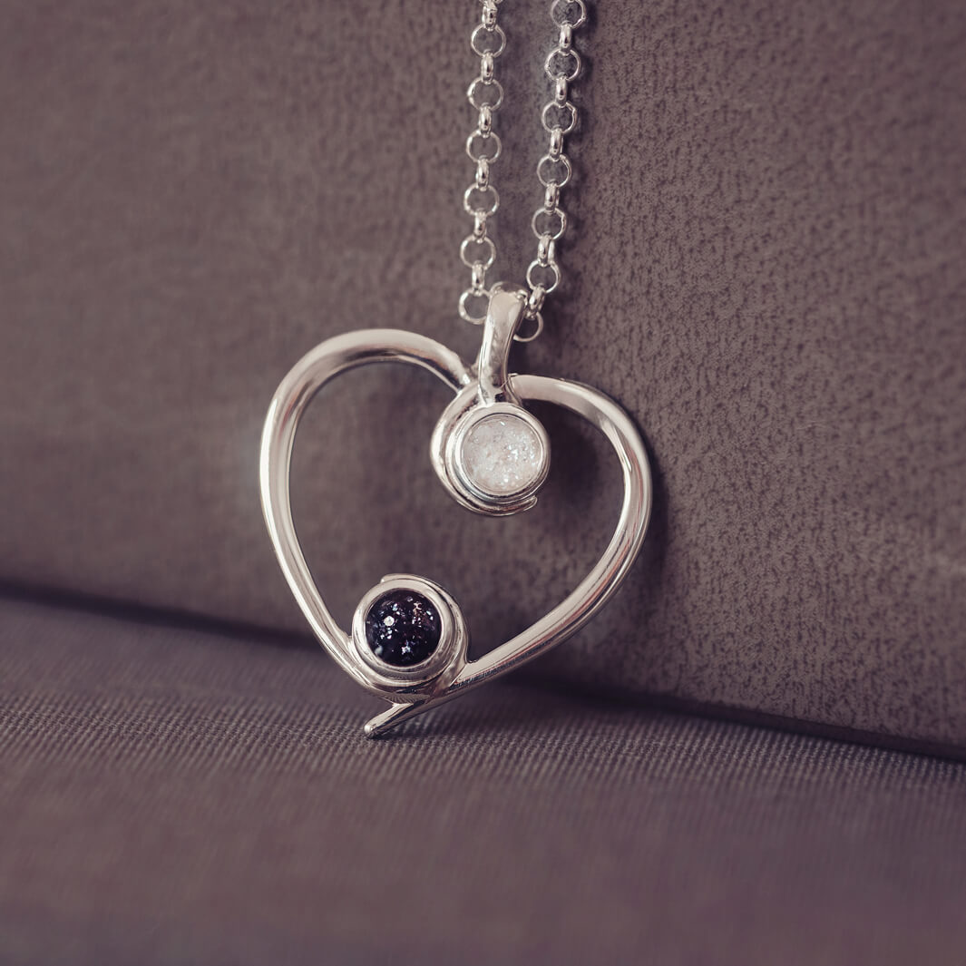 A silver heart-shaped pendant necklace featuring two sparkling black and clear resin stones, holding cremation ashes, displayed against a neutral background.
