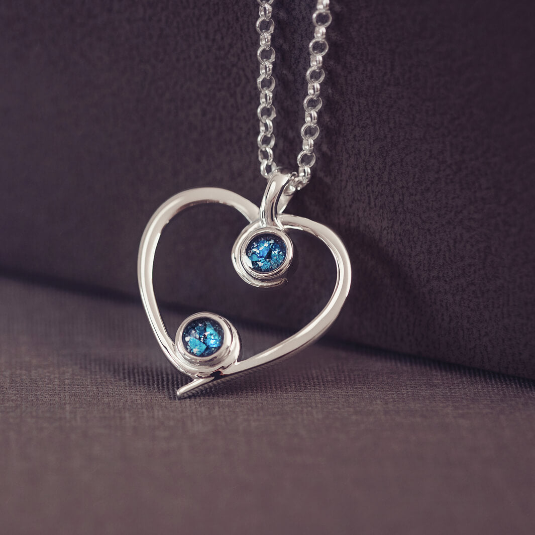 A silver heart-shaped pendant necklace featuring two sparkling blue resin stones, holding cremation ashes, displayed against a neutral background.