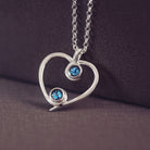 A silver heart-shaped pendant necklace featuring two sparkling blue resin stones, holding cremation ashes, displayed against a neutral background.
