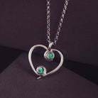 A silver heart-shaped pendant necklace featuring two sparkling green resin stones, holding cremation ashes, displayed against a dark background.