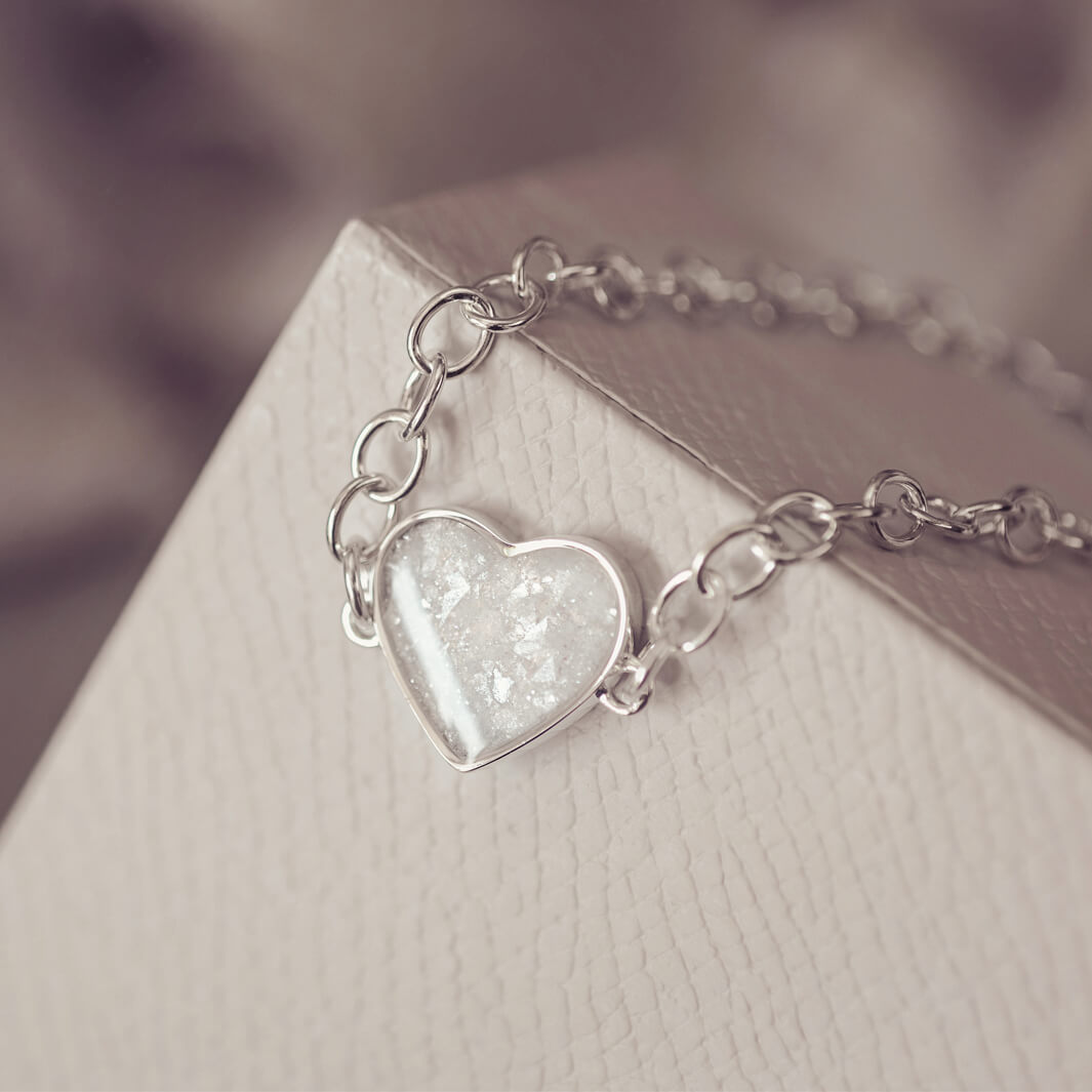 silver bracelet & heart charm, holding clear crystal, resin & cremation ashes. laying on a grey jewellery box