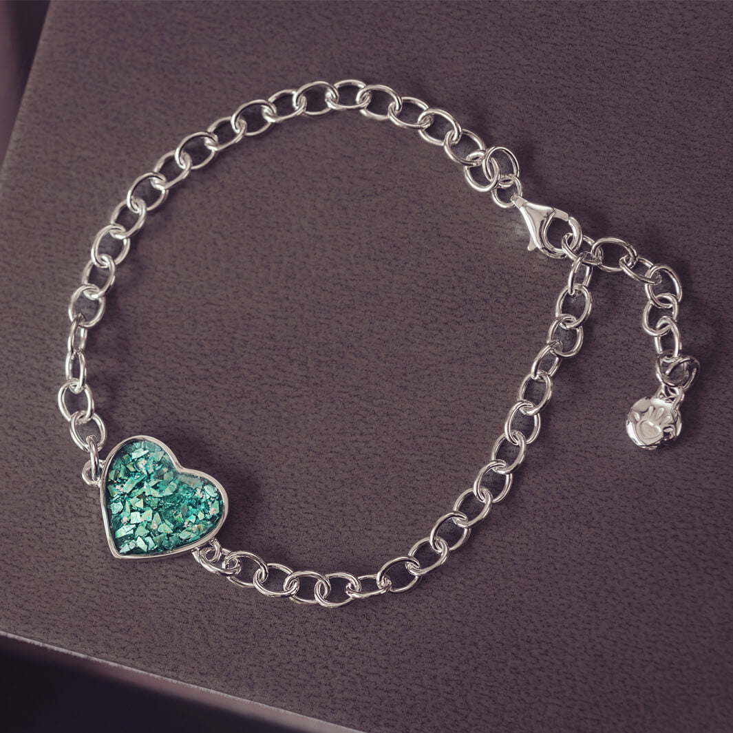 silver bracelet & heart charm, holding green crystal, resin & cremation ashes. laying on a black back ground 