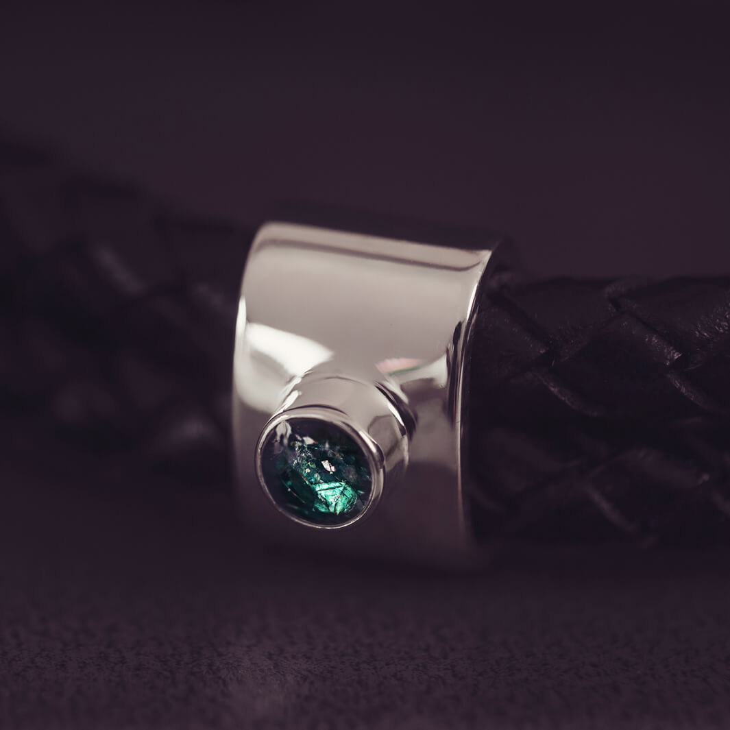 Black braided leather bracelet featuring a silver charm with a small green memorial stone set in the centre, displayed on a grey surface.