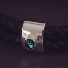 Black braided leather bracelet featuring a silver charm with a small green memorial stone set in the centre, displayed on a grey surface.