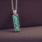 A sophisticated silver necklace featuring a rectangular pendant filled with shimmering green crystal and resin, delicately layered with cremation ashes, showcased against a dark background, on a silver belcher chain.