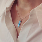 A sterling silver rectangle necklace, holding blue crystal and cremation ashes, on a silver chain being worn around a ladies neck