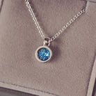 Sterling silver small round necklace with a central section filled with blue memorial ashes and crushed crystals, displayed on a beige fabric background.