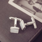 Square silver cufflinks containing cremation ashes, clear crystal and resin laying on their side on a black box
