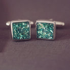 Square silver cufflinks containing cremation ashes, green crystal and resin sat on a dark grey background