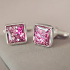 Square silver cufflinks containing cremation ashes, pink crystal and resin sat on a light grey background
