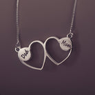 A delicate silver necklace featuring two intertwined heart-shaped pendants, engraved with a three character name on each heart. Displayed against a neutral background.