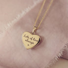 Reverse of the small gold heart necklace with engraving in our standard font laying on a beige fabric background 