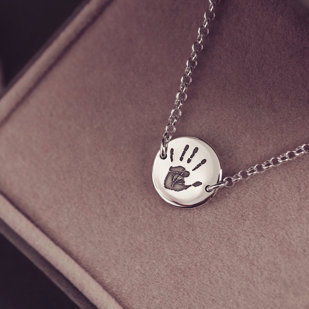 A round silver pendant bracelet featuring a detailed engraved handprint, displayed on a delicate silver chain. The necklace rests in an elegant, soft velvet jewellery box
