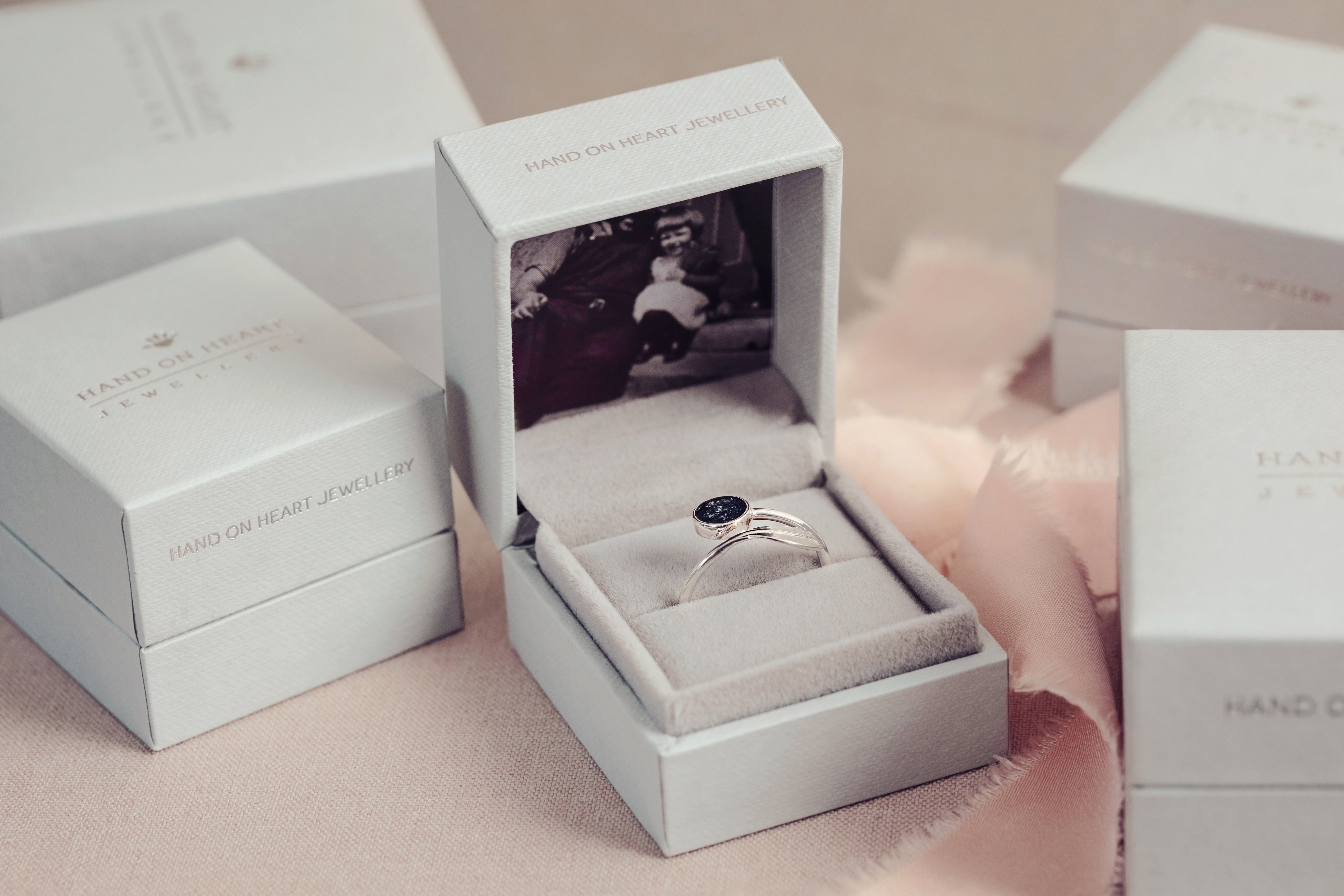 A silver ashes feather ring displayed in a soft grey jewellery box surrounded by similar jewellery boxes