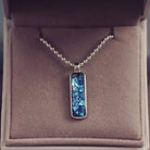 A sophisticated Mens silver necklace featuring a rectangular pendant filled with shimmering blue crystal and resin, delicately layered with cremation ashes, showcased against a dark background on a silver ball chain