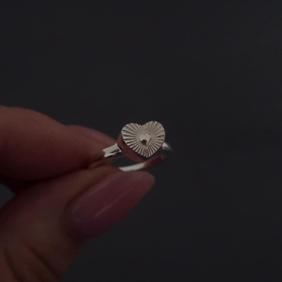 womans hands with pink nails demonstrating how to fill a silver heart ring agaisnt a dark background. It shows her unscrewing the screw and using a funnel to fill the ring with ashes. its then displayed in a grey jewellery box at the end.