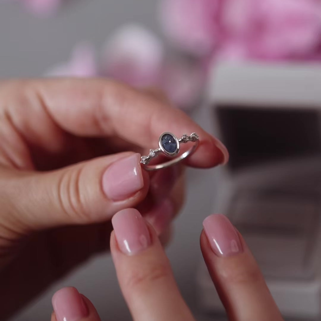 Silver ashes oval ring with blue crystals being placed onto a women's finger with pink nail polish