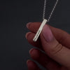 womans hands with pink nails doing a demonstration of how to fill a silver bar necklace with ashes. The necklace has "Michael" engraved on the front and "18.03.1974" on the back. it shows her using a screwdriver to remove the screw then a funnel to fill with ashes.