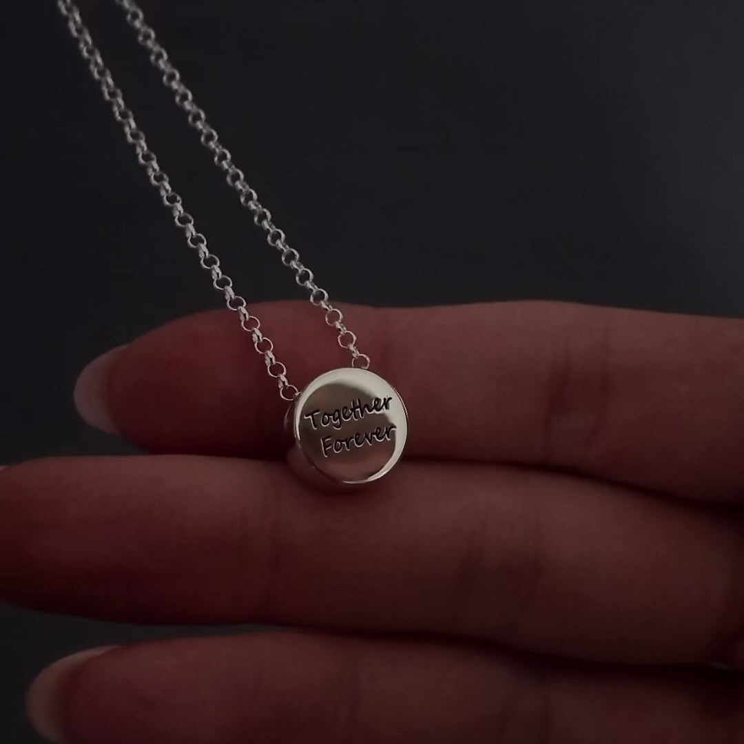 womans hands with pink nails demonstrating how to fill a silver round pendant agaisnt a dark background. It shows her unscrewing the screw and using a funnel to fill the pendant with ashes. its then displayed in a grey jewellery box at the end.