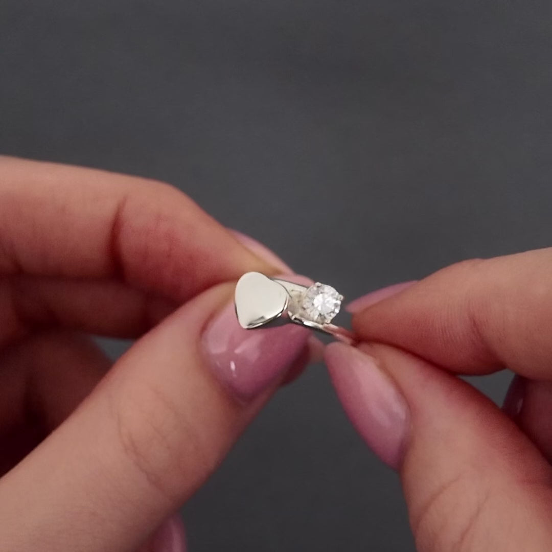 womans hands holding a silver wrap heart ring. the video shows a demonstration of filling the ring with ashes by unscrewing the screw with a screwdriver and using a funnel to scoop ashes into the ring.