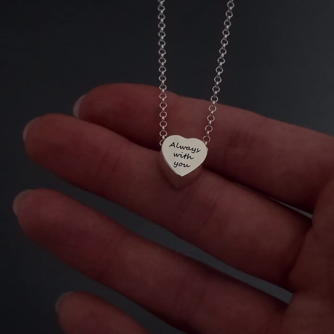 womans hands with pink nails demonstrating how to fill a silver heart pendant agaisnt a dark background. It shows her unscrewing the screw and using a funnel to fill the pendant with ashes. its then displayed in a grey jewellery box at the end.