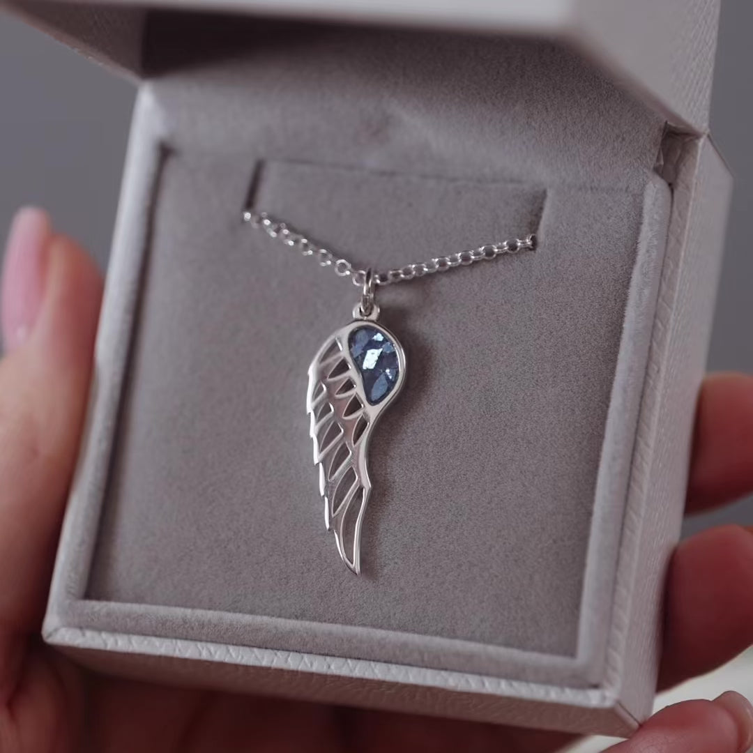 Silver ashes jewellery wing pendant with blue crystals in a grey jewellery box held in a women's hand.