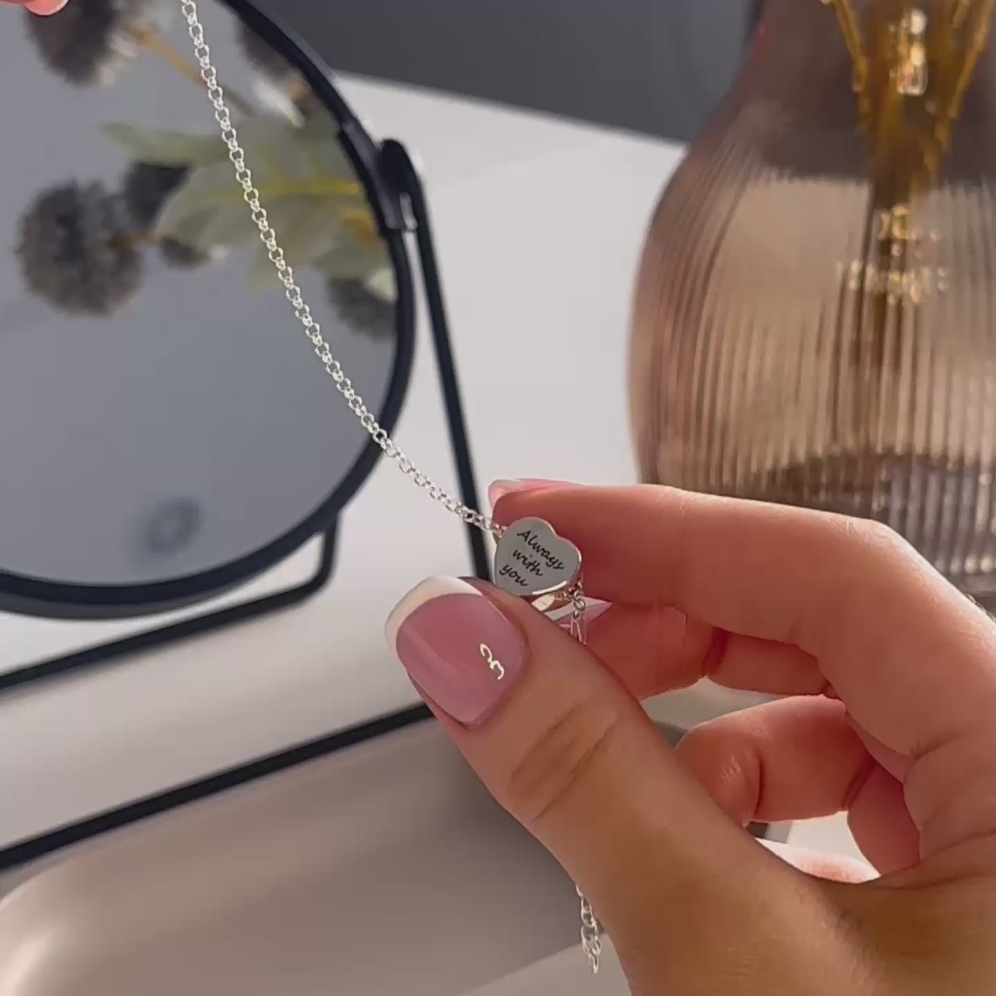 video showing a demonstration of filling an ashes urn necklace. the necklace is a silver heart pendant with "always with you" written on the front. the woman's hands show unscrewing the pendant and using a stick to place to ashes inside. the woman has pink nails with white tips. there is a black mirror, brown flower vase and a grey trinket dish n the background.