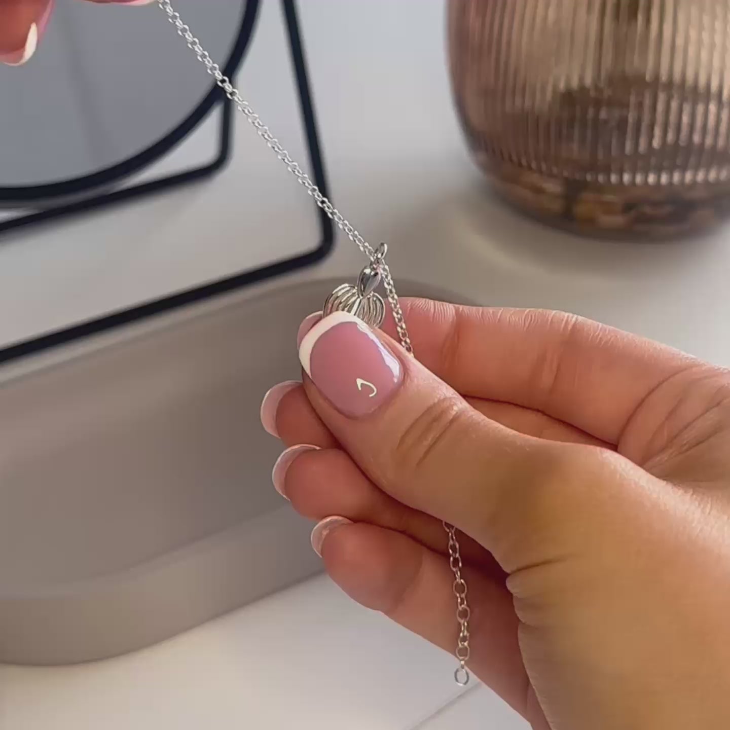 video showing a demonstration of a woman's filling a silver angel necklace with cremation ashes. The woman has pink nails with white tips. there is a black mirror, brown flower vase and a grey trinket dish in the background.