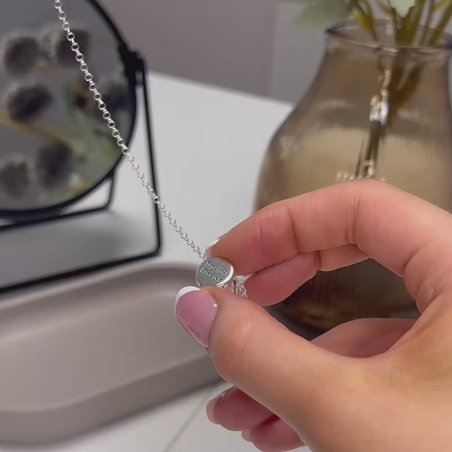 demonstration video showing a woman's hands filling an ashes urn necklace. the urn necklace is a round silver pendant with "together forever" written on the front. the video shows the woman's hands unscrewing the pendant then filling it with ashes. the woman has pink nails with white tips. there is a black mirror, brown flower vase and a grey trinket dish in the background.