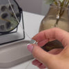 demonstration video showing a woman's hands filling an ashes urn necklace. the urn necklace is a round silver pendant with "together forever" written on the front. the video shows the woman's hands unscrewing the pendant then filling it with ashes. the woman has pink nails with white tips. there is a black mirror, brown flower vase and a grey trinket dish in the background.
