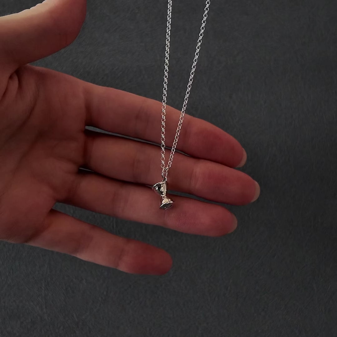 Demo video of womans hands with pink nails showing how to fill a silver hourglass urn necklace pendant. it shows her unscrewing the bottom of the pendant with a screwdriver and using a funnel to fill the hourglass with cremation ashes. the screw is then screwed back on and the necklace is displayed in a grey jewellery box. 